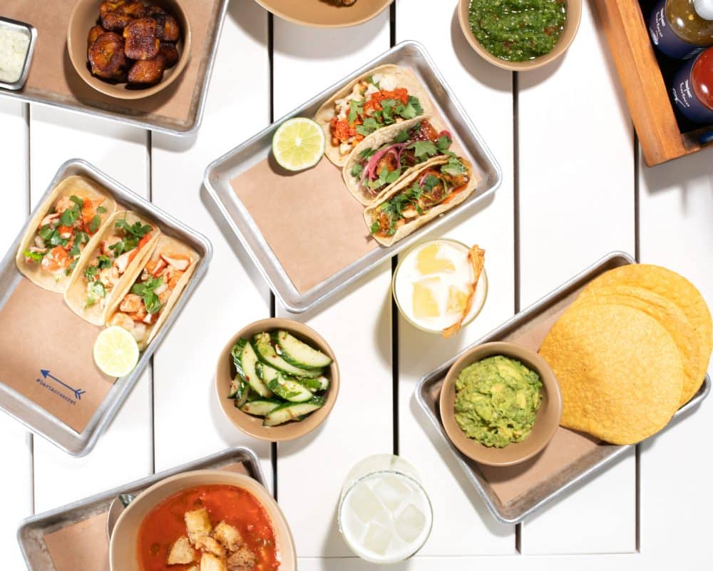 table with spread of bright + colorful food including trays of tacos, gazpacho, chips + guac, cucumber salad, plantains and cocktails