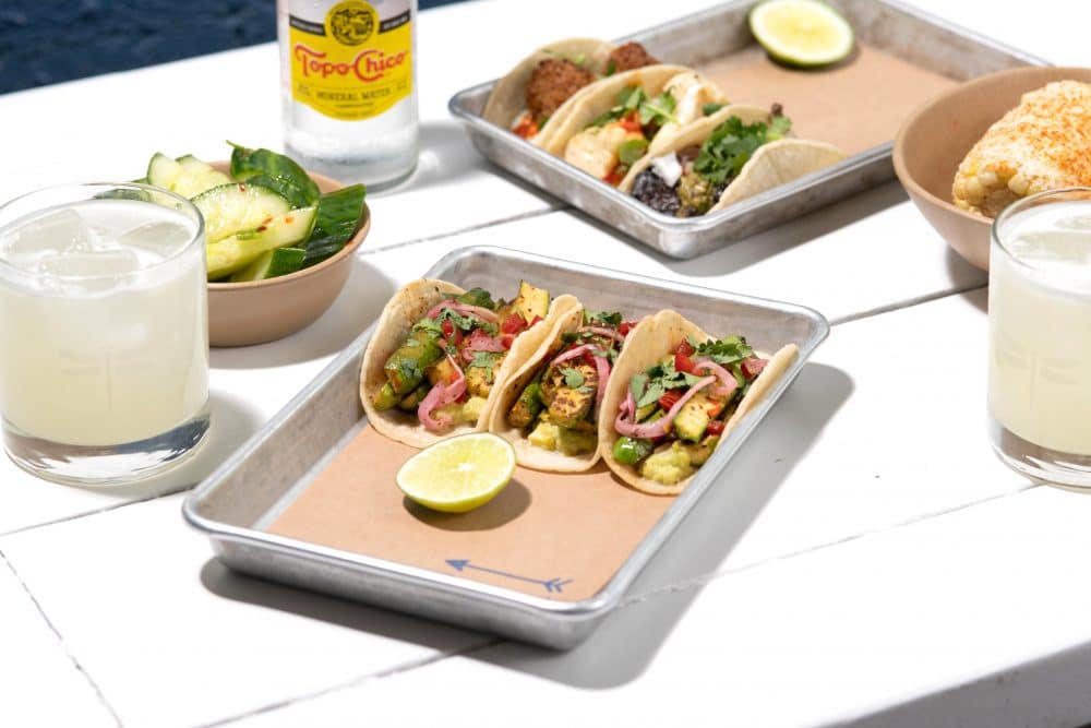 image of a table with a spread of food including tray with three #bartacosecret baby zucchini tacos, cocktails, and another tray of a variety of tacos