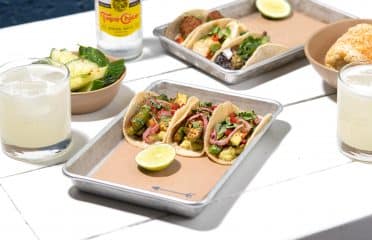 image of a table with a spread of food including tray with three #bartacosecret baby zucchini tacos, cocktails, and another tray of a variety of tacos