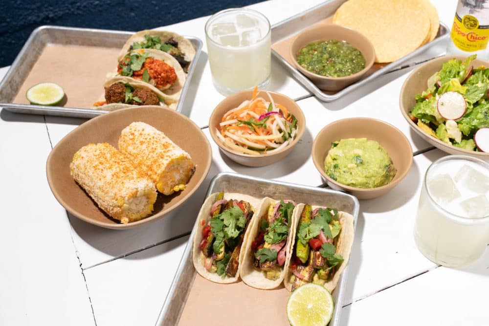 image of table with a spread of bright food including a tray with three #bartacosecret baby zucchini tacos, street corn, papaya salad, guacamole, chips and salsa verde, and two margaritas.