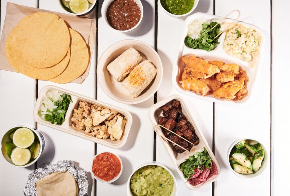 image of spread of take out food including bartaco taco packs with glazed pork belly, baja fish and cauliflower tacos, plus street corn, spicy cucumber salad, and chips + guac + salsa